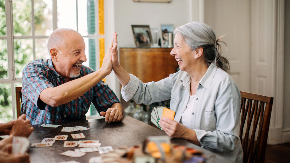 Ein älterer Mann und eine ältere Frau spielen an einem Tisch ein Kartenspiel und machen lachend ein High Five.